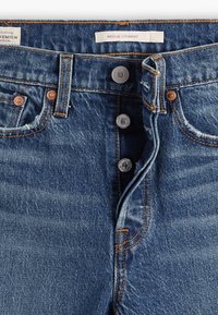 Jeans de denim en azul oscuro con un corte recto. Presentan cinco botones metálicos, remaches de cobre y costuras naranjas en contraste. La etiqueta dice "Wedgie Straight."