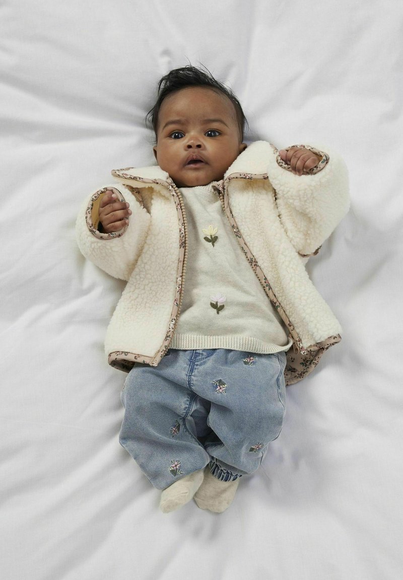 Texturerad krämfärgad fleecejacka med blommönster, lätt tröja med broderade blommor, denimbyxor med blommönster, ljus bakgrund.