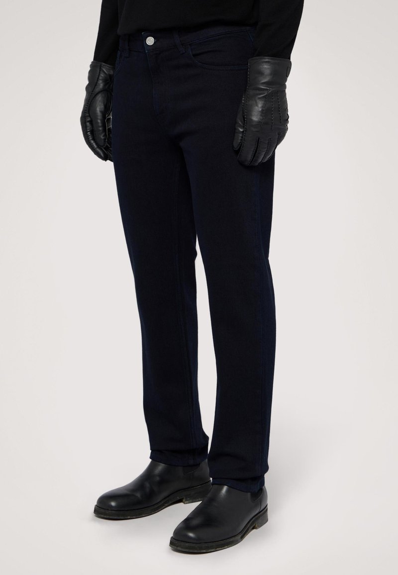 Jeans azul marino de denim, con un ajuste entallado, bolsillos frontales, cierre de botón metálico, y combinados con guantes y botas de piel negra.