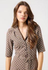 Vestito marrone con motivo, dotato di maniche corte, colletto e cintura annodata in vita. Il tessuto ha una sottile texture con accenti bianchi a contrasto.
