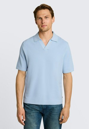 Mann steht und trägt ein hellblaues kurzärmeliges Strick-Polohemd und blaue Jeans vor einem schlichten hellen Hintergrund.