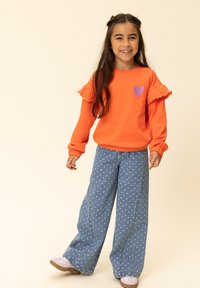 Oranje sweatshirt met lange mouwen en ruches aan de mouwen, met een paarse hartenpatch, gecombineerd met wijde blauwe spijkerbroek met witte hartjes.