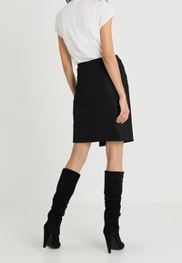 Zwarte pencilrok met een gestructureerde afwerking, gecombineerd met een wit topje met korte mouwen. Het model draagt zwarte suede knie-hoge stiletto laarzen.