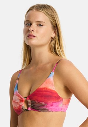 Kobieta z długimi blond włosami w kolorowym, farbowanym bikini z centralnym pierścieniem, stojąca na jednolitym tle.
