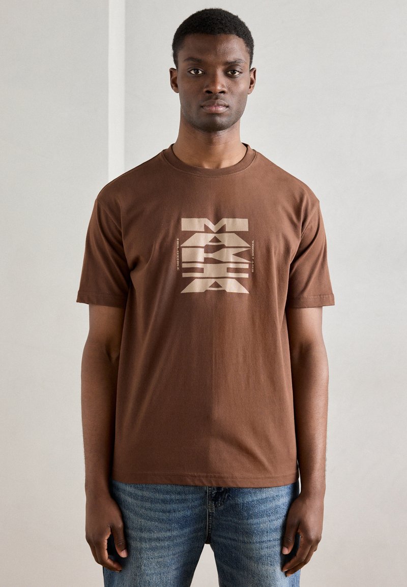 Brun bomulds t-shirt med et grafisk design i beige, der har abstrakte former og tekst. Korte ærmer og en klassisk rund halsudskæring.