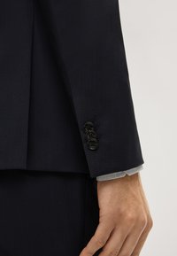 Dunkelblaues Sakkos mit drei schwarzen Knöpfen und gewebter Textur, aus dem der Ärmel eines grau-weißen karierten Hemdes herausblitzt.