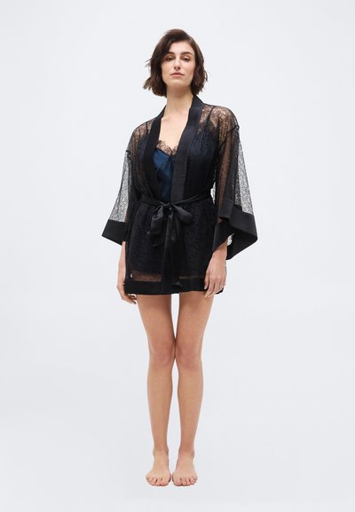 Mujer de pie descalza, vistiendo una bata transparente de encaje negro con ribete de satén y cinturón atado sobre lencería azul oscuro.