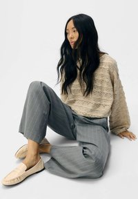 Beige gestrickter Pullover mit strukturiertem Muster, graue gestreifte weite Hose und beiger Slipper, vor einem schlichten weißen Hintergrund.