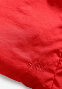 Červená tkanina s hladkým povrchom, s vyšitým logom "self." v mierne tmavšej farbe, umiestneným v pravom dolnom rohu.