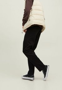 Beżowa kamizelka puchowa na brązowym swetrze, w połączeniu z czarnymi spodniami i czarnymi trampkami za kostkę z białymi podeszwami i sznurowadłami.