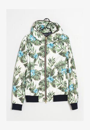 Biała kurtka z kapturem na suwak, w zielony tropikalny wzór z liści i niebieskimi kwiatami, z czarnymi ściągaczami na rękawach i w pasie, wisząca na wieszaku.