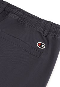 Navy elastische broek met een zijzak, voorzien van een wit en rood geborduurd logo aan de zijkant en een gladde, moderne textuur.