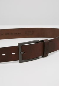 Brauner Ledergürtel mit einer matt schwarzen Metallschnalle, die eine strukturierte Oberfläche und mehrere Größenlöcher zur Anpassung aufweist. Eingravierte Details auf dem Leder.