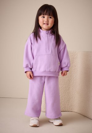 Ung pige står iført en lila sweatshirt og matchende vidde bukser med hvide sneakers mod en neutral baggrund.