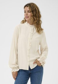 Femme aux cheveux ondulés portant un blouse crémeuse à volants avec des boutons et un jean bleu, se tenant avec une main dans sa poche, regardant sur le côté.