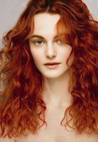Modelo con abundante cabello rizado y rojizo alrededor de la cara, piel clara y ojos azul claro, mostrando un maquillaje natural y minimalista.