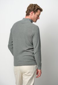 Grau gestrickter Pullover mit hohem Kragen, strukturiertem Stoff und langen Ärmeln, kombiniert mit hellen Hosen, von hinten betrachtet.