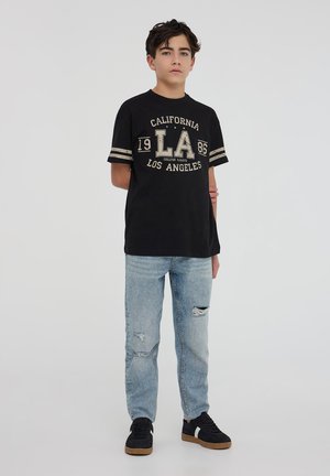 T-shirt nera con scritte dorate, con "CALIFORNIA" e "LA 1986." Abbinata a jeans chiari strappati e sneakers nere.