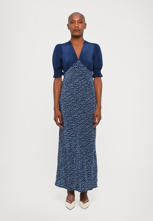 Navyblauw jurk met pofmouwen en een V-hals. Bevat een stippenpatroon en heeft een getailleerde pasvorm die uitloopt aan de onderkant. Draagt met witte hakken.