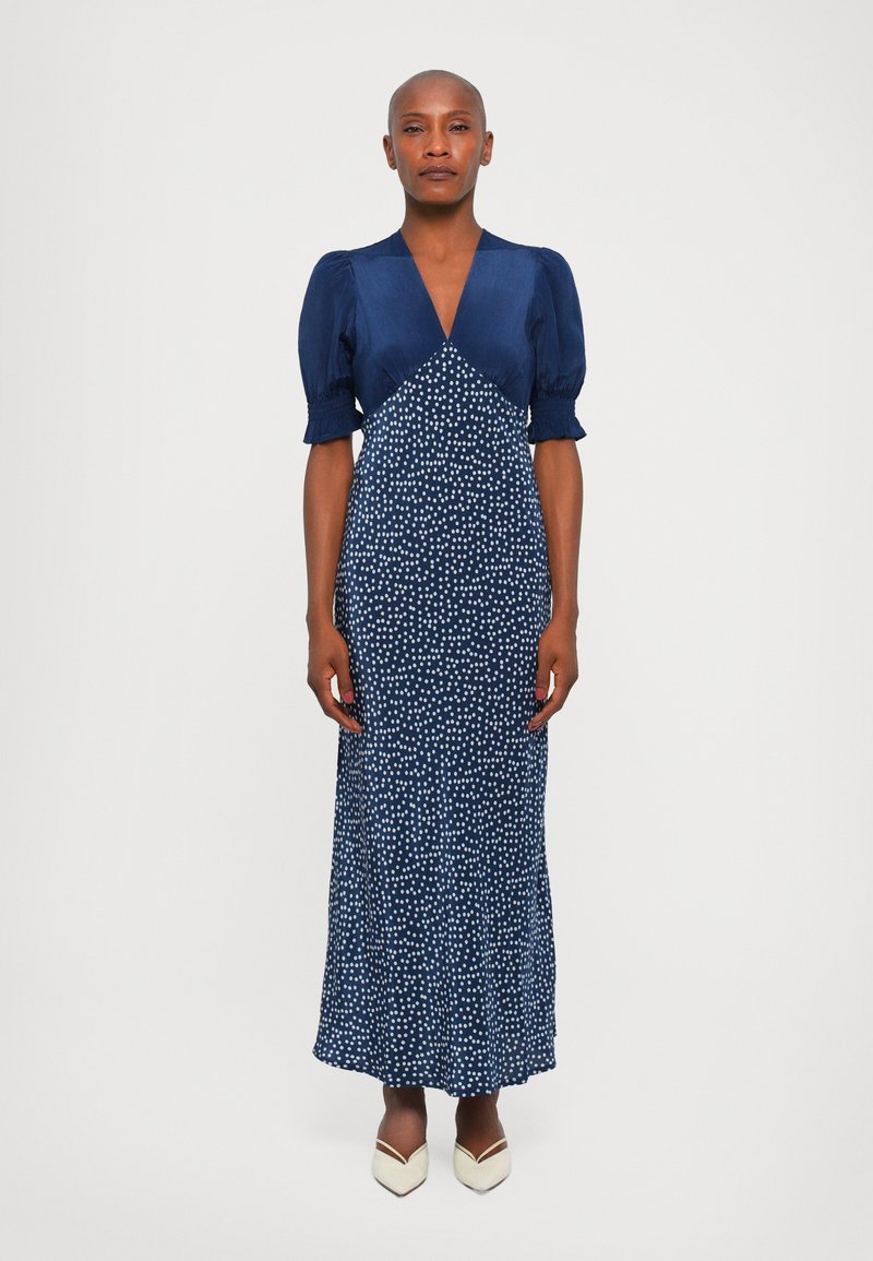 Navyblauw jurk met pofmouwen en een V-hals. Bevat een stippenpatroon en heeft een getailleerde pasvorm die uitloopt aan de onderkant. Draagt met witte hakken.