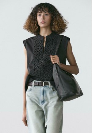 Femme aux cheveux bouclés portant un haut noir texturé sans manches, un jean en denim clair, une ceinture noire, et tenant un grand sac en cuir noir.