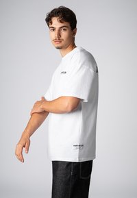 Weißes Baumwoll-T-Shirt mit kurzen Ärmeln, ausgestattet mit einem Logo auf der Vorder- und Rückseite. Kombiniert mit dunkelblauen Jeans. Einfaches Design.