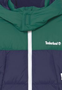 Prošívaná bunda v zelené a námořnické modři, s zapínáním na zip, kapucí se stahovací šňůrkou a logem Timberland. Hladká, lehce lesklá tkanina.