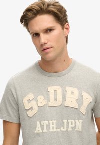 Grijze katoenen t-shirt met textuur en crème-kleurige letters "S&DYW ATH.JPN" over de borst. Ronde halslijn en korte mouwen.