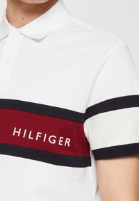 Valkoinen polo paita, jossa on punaisia ja tummansinisiä raitoja ja "HILFIGER" brodeerattu valkoisella. Kauluksellinen, nappiextra, puuvillamateriaalia.