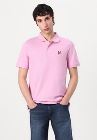 Rosa Baumwollpolohemd mit Kragen, kurzen Ärmeln und einem gelben Schmetterlingslogo auf der linken Brust. Getragen mit dunkelblauen Jeans.