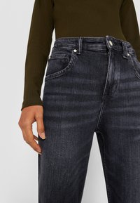 Vaqueros de tiro alto en gris oscuro con un diseño clásico de cinco bolsillos, con herrajes plateados y costuras visibles, combinados con una camiseta verde de canalé.