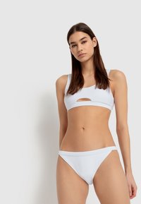 Baltas bikinis, sudarytas iš sportinio stiliaus viršaus su išpjova ir atitinkančiais žemais subtiliais apatiniais, pagamintas iš tamprios ir glotnios medžiagos.