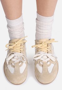 Feet wearing white ribbed socks and beige sneakers with white and yellow laces tied in bows on a white background.