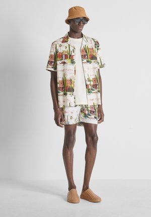 Hombre con camisa de manga corta y pantalones cortos a juego con estampado de cactus y desierto, sombrero tipo bucket color tostado, gafas de sol, zapatos beige sin cordones.