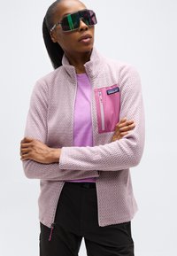 Γυναίκα που φοράει υπερμέγεθες αντανακλαστικά γυαλιά ηλίου, υφασμάτινο μπουφάν ανοιχτού μοβ με ροζ τσέπη στο στήθος και μαύρο παντελόνι, με σταυρωμένα τα χέρια.