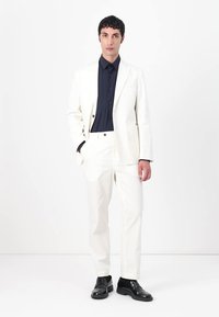 Witte op maat gemaakte pak met een geknipte revers, een enkelvoudig knoopschema en rechte broek, gecombineerd met een donkerblauw overhemd met knopen en zwarte schoenen.