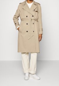 Beige trenchcoat, jossa on kaksirivinen etuosa, mustat napit, vyö ja olkapeitteet, yhdistetty valkoisiin housuihin ja lenkkareihin.