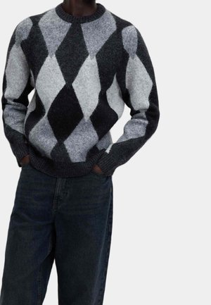 Mand iført en fuzzy strikket sweater med stort sort, gråt og hvidt argyle-mønster og mørkeblå jeans, hænder i lommerne.