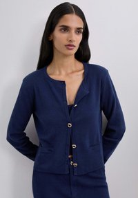 Mujer con cabello largo y oscuro, vestida con un cárdigan azul marino con cierres de botones dorados y pantalones a juego, de pie contra una pared clara y sencilla.