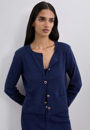 Mujer con cabello largo y oscuro, vestida con un cárdigan azul marino con cierres de botones dorados y pantalones a juego, de pie contra una pared clara y sencilla.