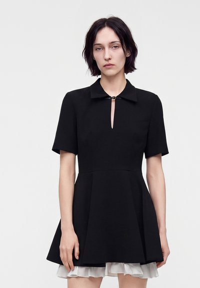 Femme aux cheveux courts et foncés portant une robe noire à manches courtes avec un petit col et un ourlet blanc froncé sur un fond uni.