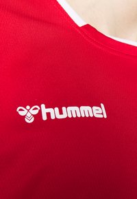 Logo Hummel blanc et texte sur un maillot de sport rouge, montrant la partie supérieure de la poitrine près de l'encolure.