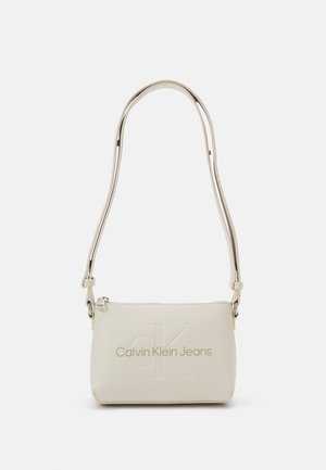 Mała beżowa torba na ramię z regulowanym paskiem, zapięciem na zamek i wytłoczonym logo Calvin Klein Jeans z przodu.