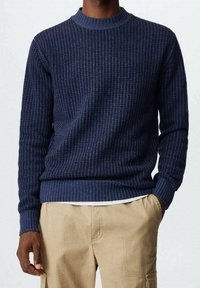 Maglione a coste blu navy con scollo rotondo e maniche lunghe, caratterizzato da un motivo testurizzato e un'orlo aderente. Indossato sopra pantaloni di colore chiaro.