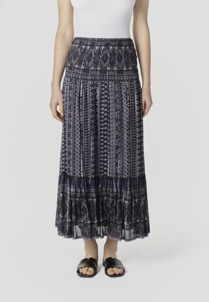 Naine, kes kannab pikka, mustrilist tumesinist ja halli seelikut, valget varrukateta särk ja musti libisevaid sandaale.