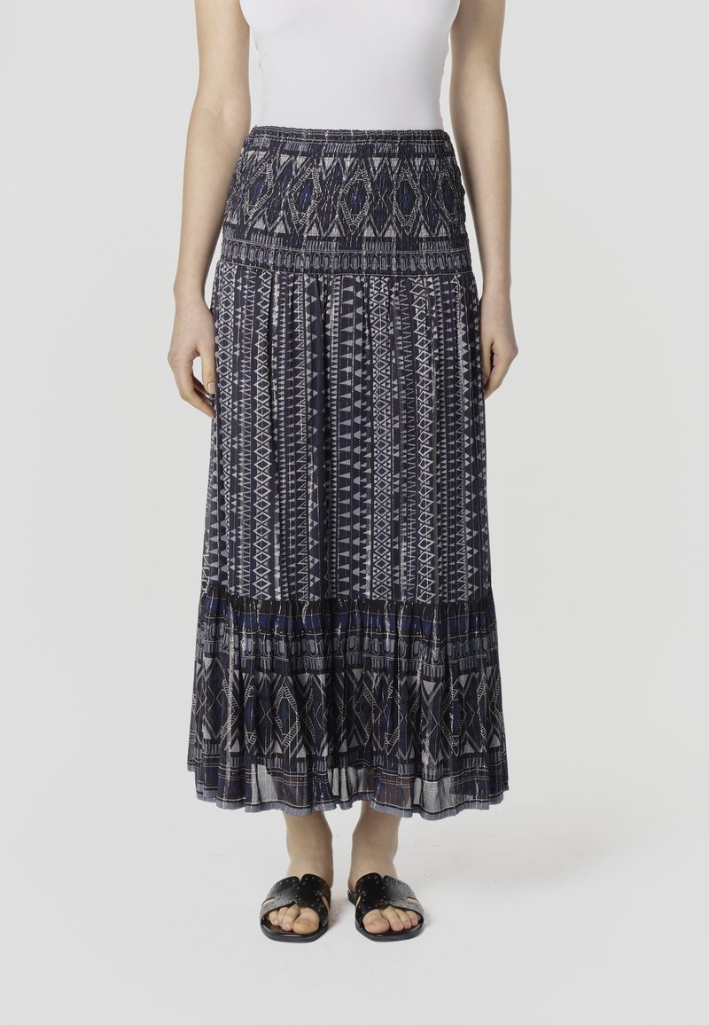 Nő hosszú, mintás tengerészkék és szürke szoknyában, fehér ujjatlan felsőben és fekete papucsban.