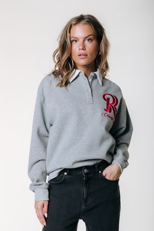 Hanorac gri cu guler alb, logo roșu „R” brodat pe piept și mâneci lungi. Purtați cu blugi negri.