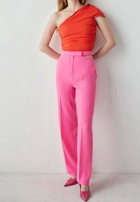 Frau trägt ein leuchtend orangefarbenes Top mit einem Schulterträger, kombiniert mit hoch taillierten rosa Hosen und rosa spitzen Schuhen, stehend vor einer einfarbigen Wand.