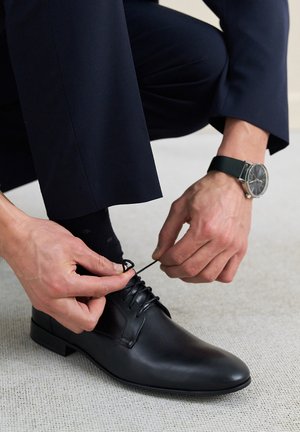 Czarne skórzane buty na dress z zaokrąglonym noskiem, z zawiązywaniem i gładkim wykończeniem. Ręce związujące sznurówki; widoczne ciemne skarpetki i zegarek.