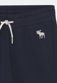 Marineblauwe trainingsbroek met elastische tailleband, witte trekkoord en een klein wit geborduurd oudste logootje op de rechterzijzak.